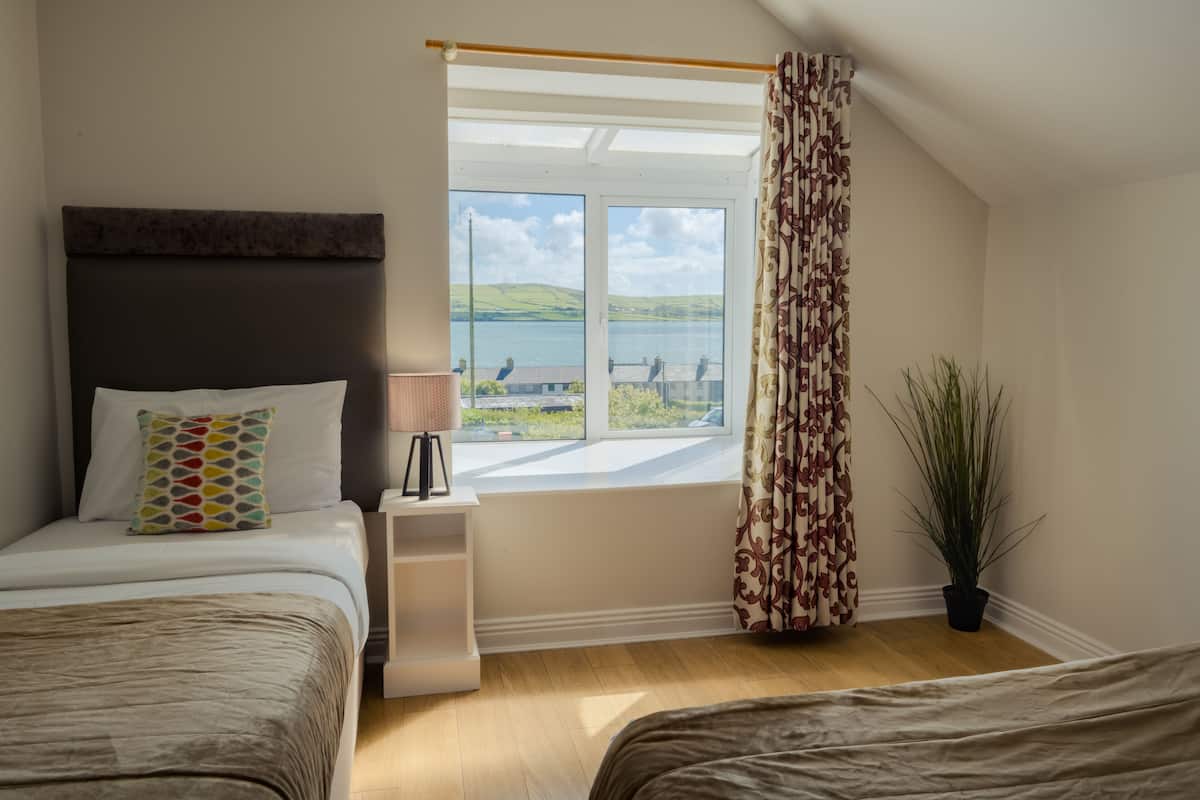 A bedroom with two beds, a window with a view of hills and water, a bedside table with a lamp, and a potted plant.