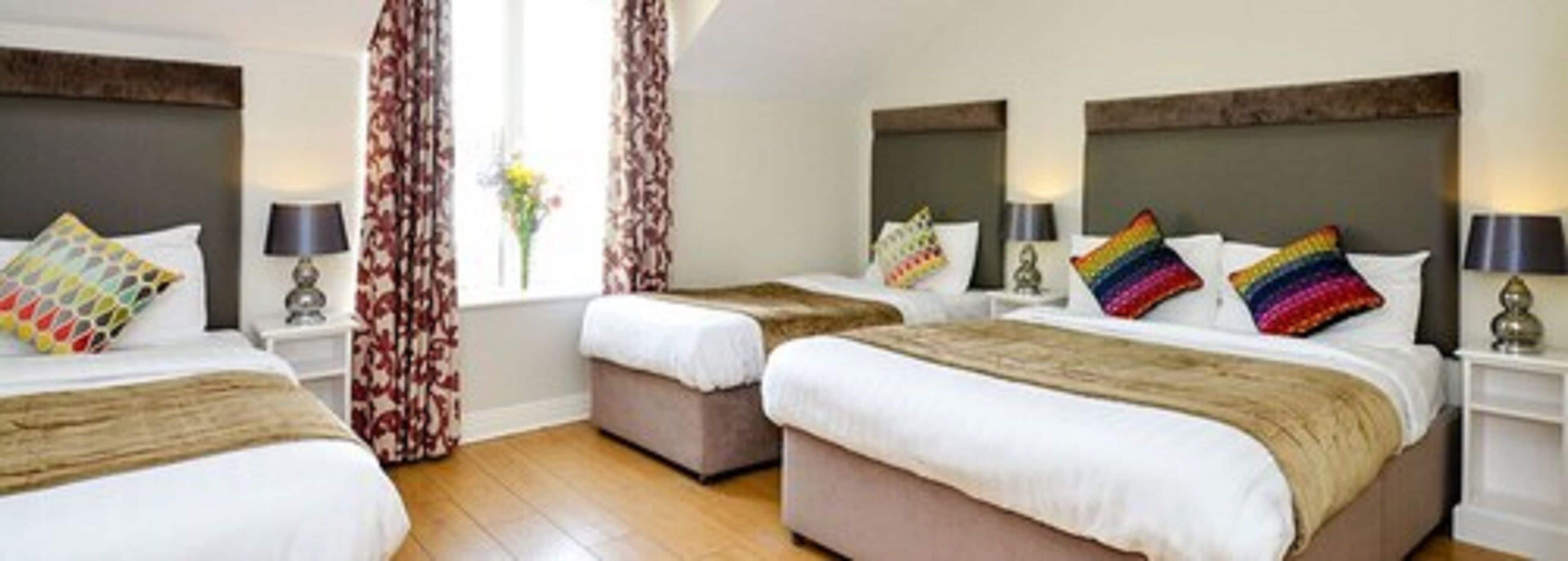 Habitación de hotel con tres camas, cada una con un cojín de estampado diferente, suelo de madera y una ventana con cortinas.