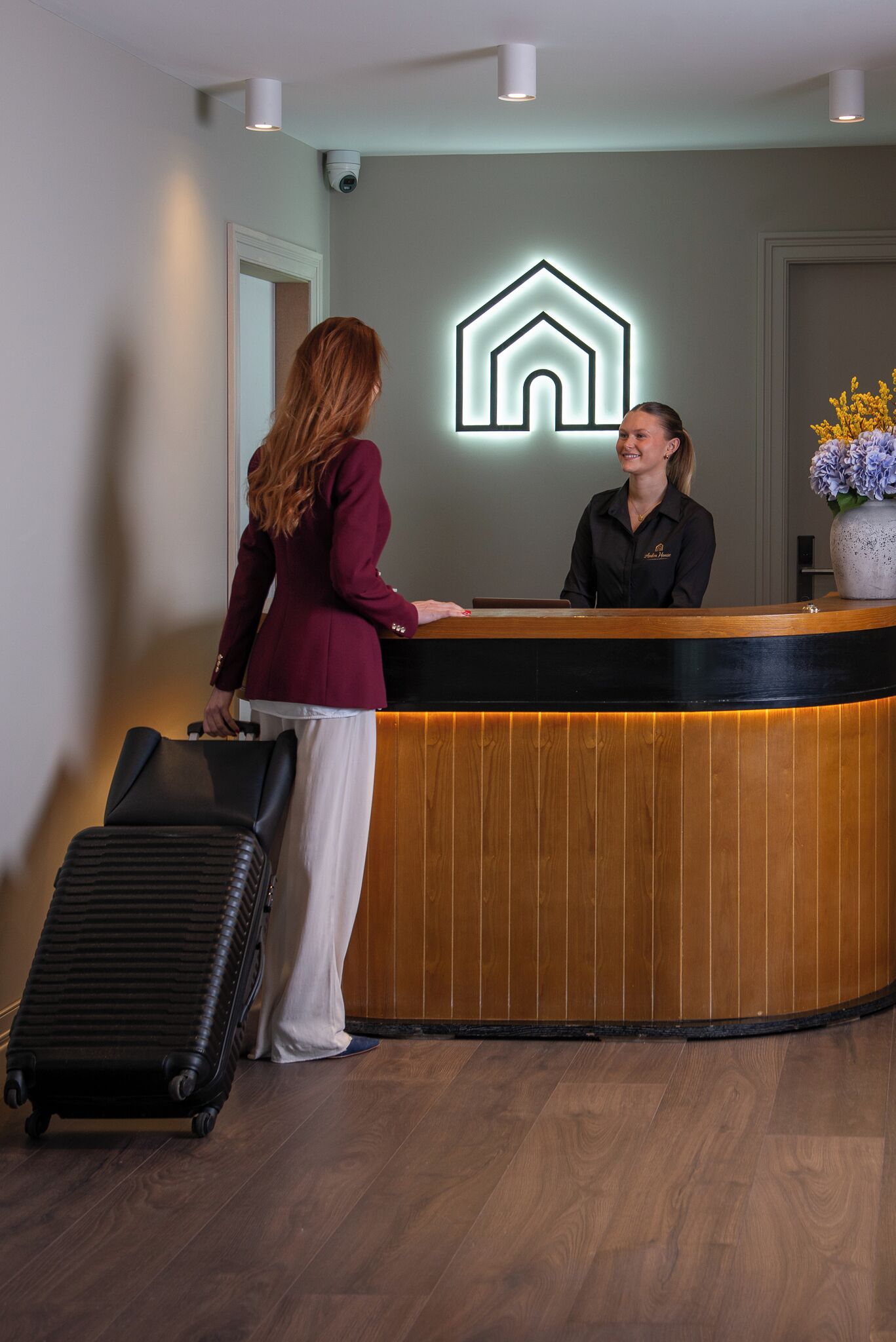 A reception desk with a wooden finish, a neon house symbol, and a potted plant.