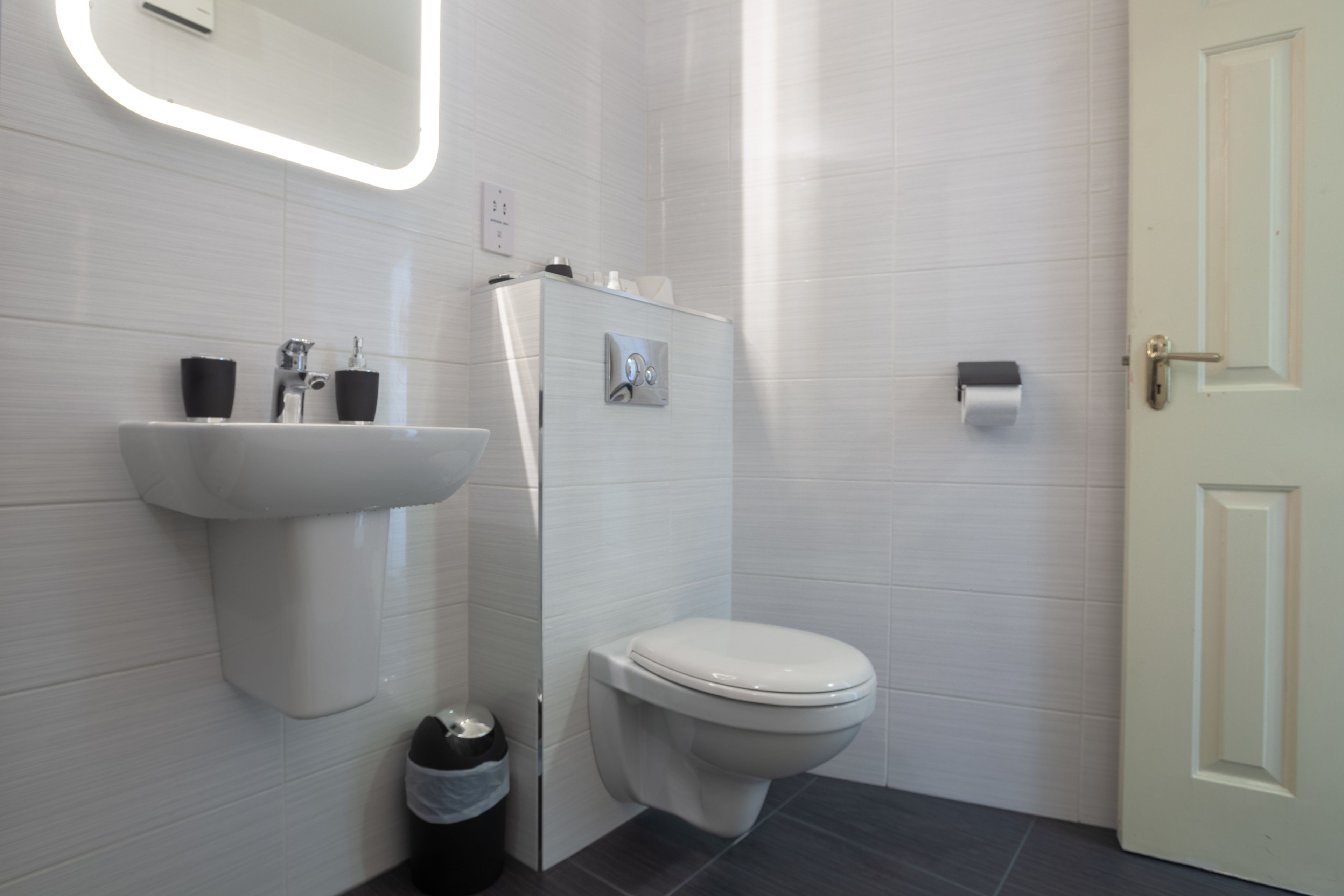 A modern bathroom with a white sink, toilet, and a mirror with built-in lighting.