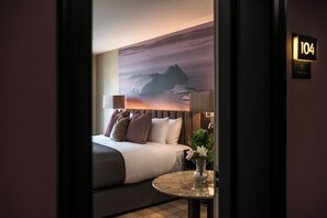 A hotel room with a bed, a round table with a vase, a wall-mounted lamp, and a large scenic painting. - Arden House (Dingle)