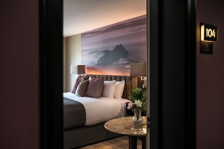 A hotel room with a bed, a round table with a vase, and a wall-mounted lamp.. Arden House