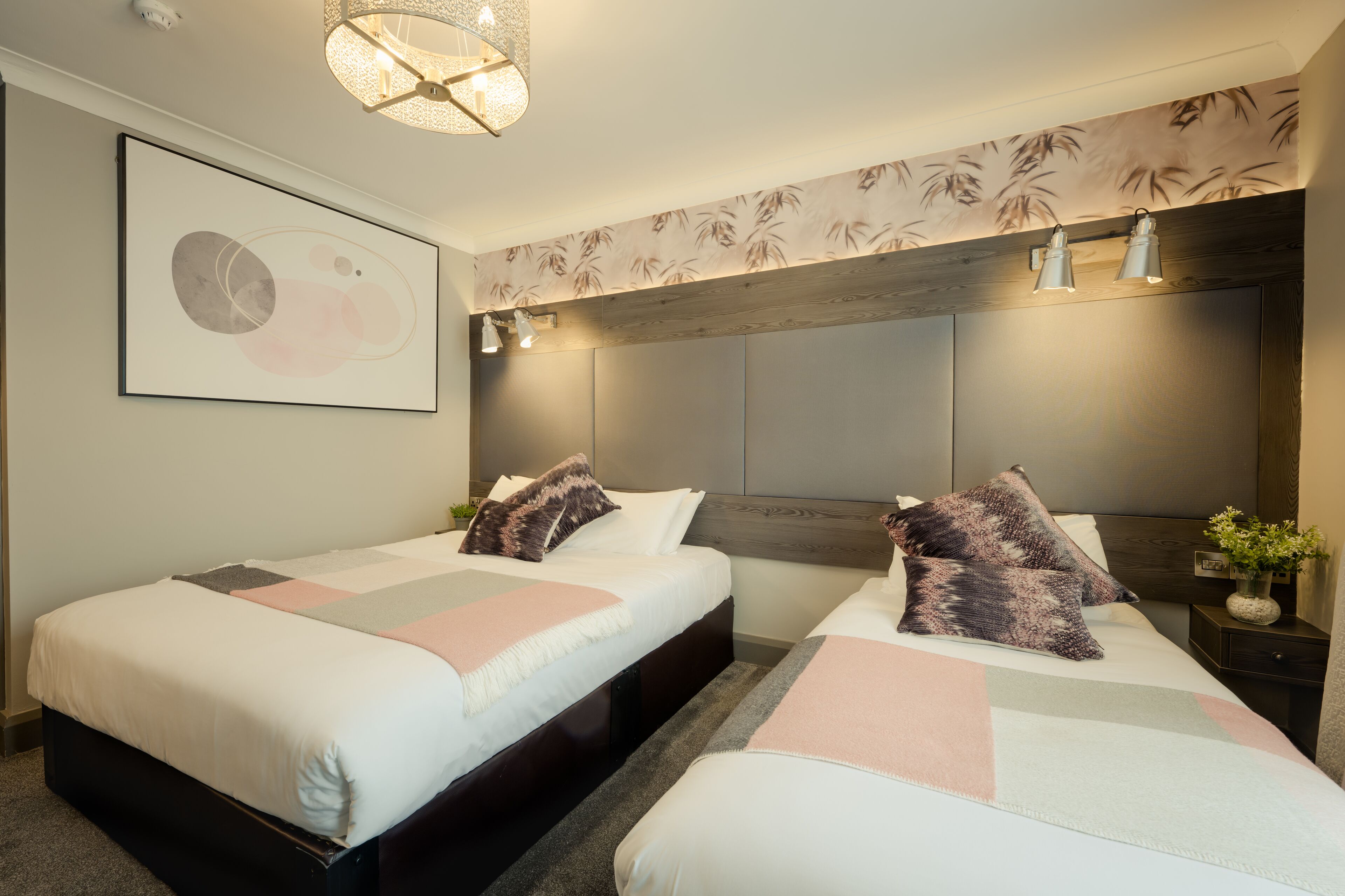 Habitación de hotel con dos camas, un cabecero moderno, una obra de arte colgada en la pared y una mesita de noche pequeña con una planta.