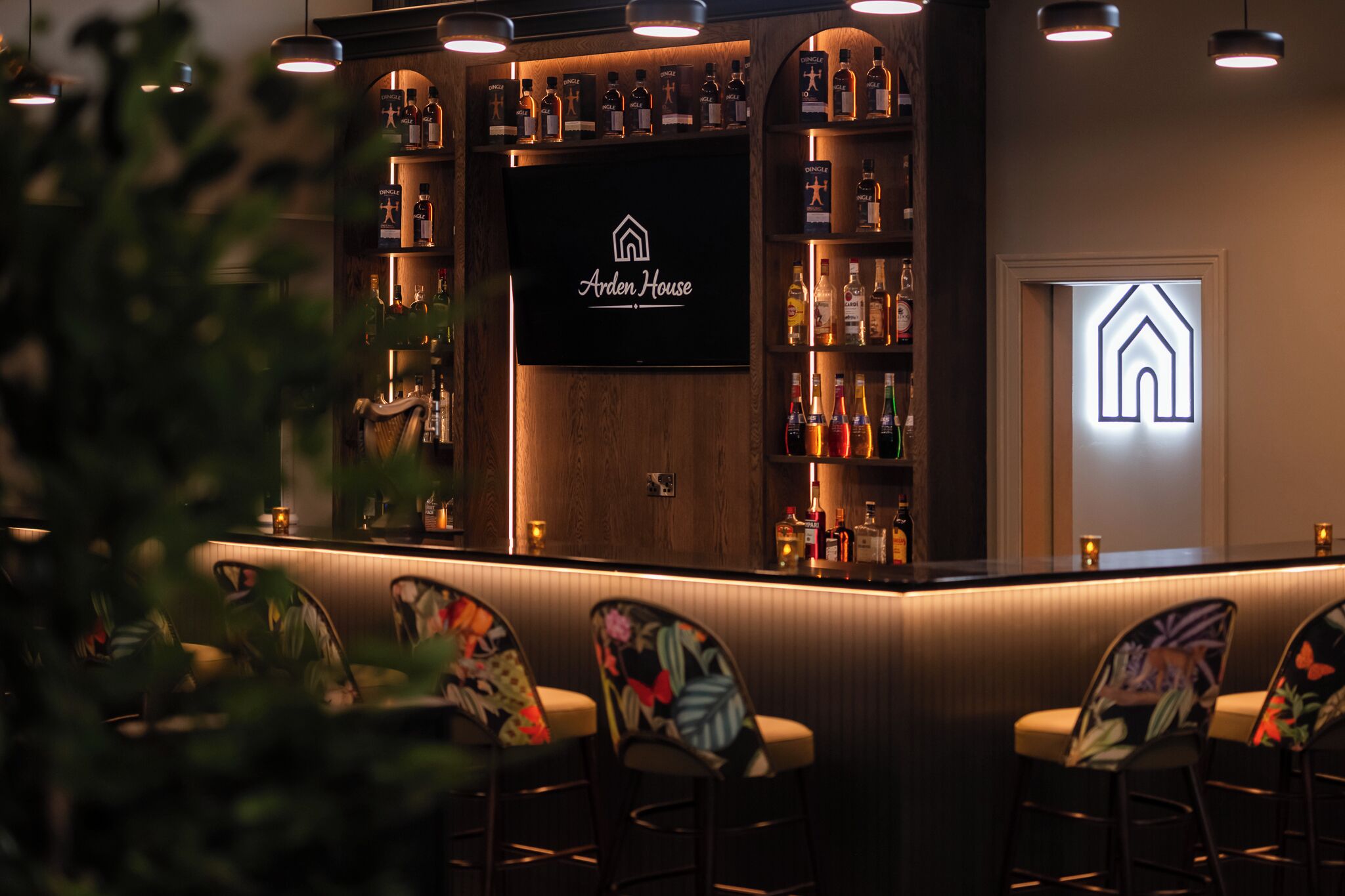 Zona de bar con una pantalla de televisión que muestra 'Arden House', estantes con botellas de licores y taburetes de bar.