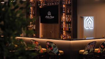 Zona de bar con una pantalla de televisión que muestra 'Arden House', estantes con botellas de licores y taburetes de bar.