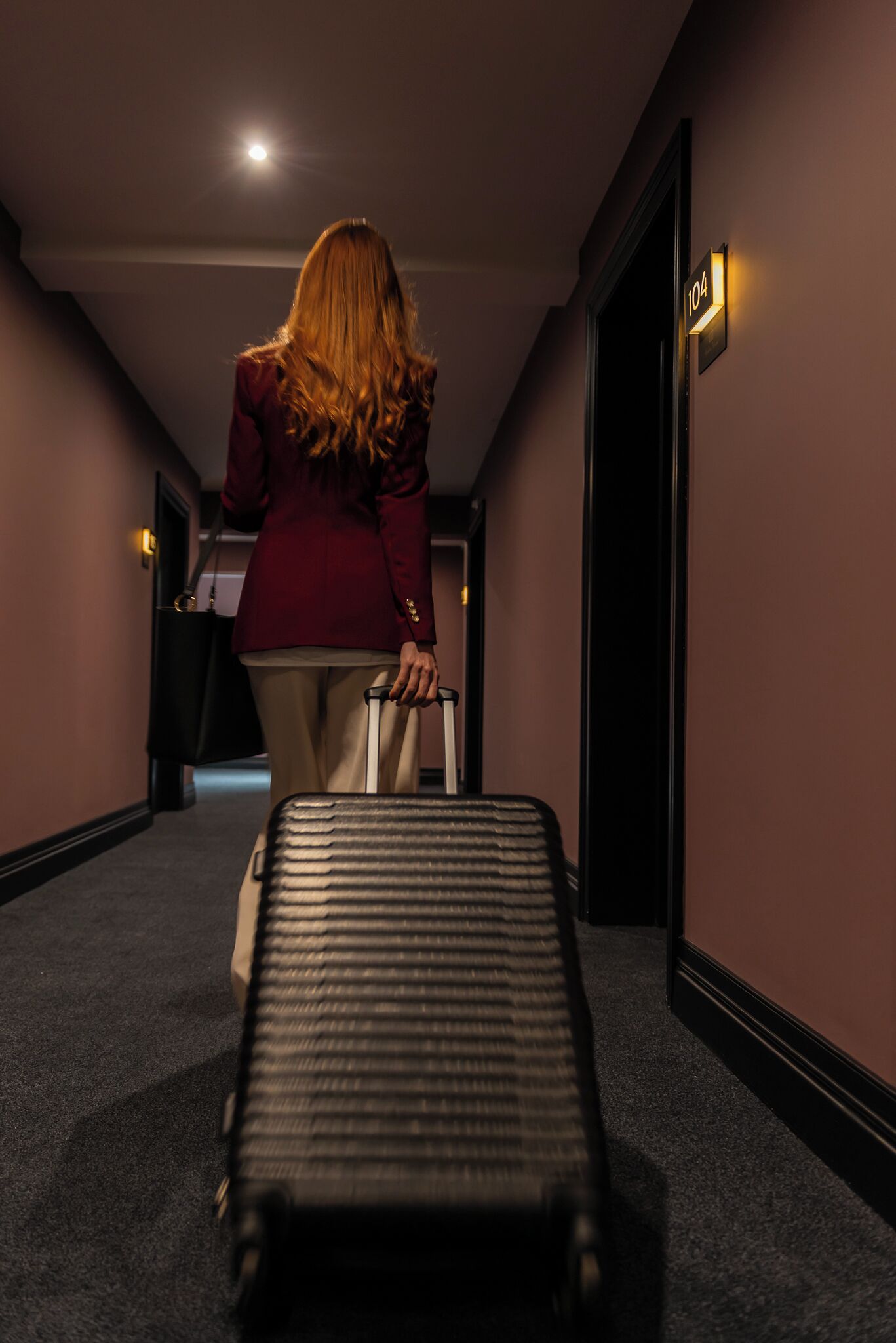 A person walking down a hallway with a suitcase, a door with a number sign, and a light fixture on the wall.