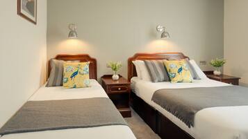 A hotel room with two beds, each with a wooden headboard, white bedding, and decorative pillows. There are bedside tables with lamps and a small plant on one of them.