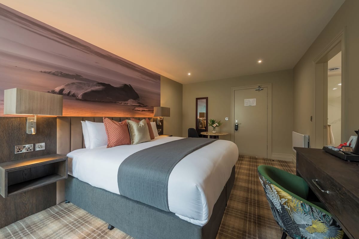 Habitación de hotel con una cama grande, un escritorio, una silla, un espejo y vistas a las montañas.