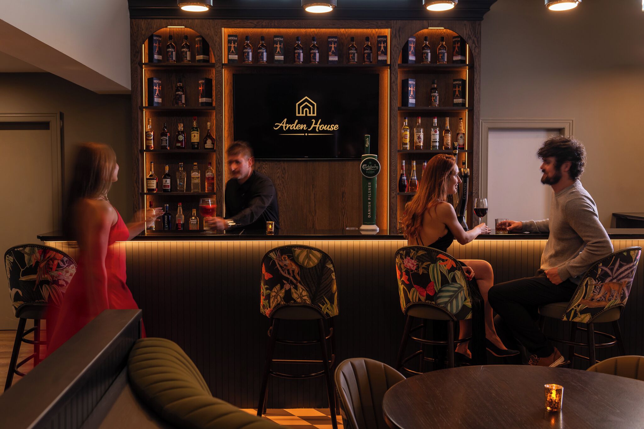 Una zona de bar con mostrador de madera, estantes con botellas y un cartel que dice 'Arden House'.