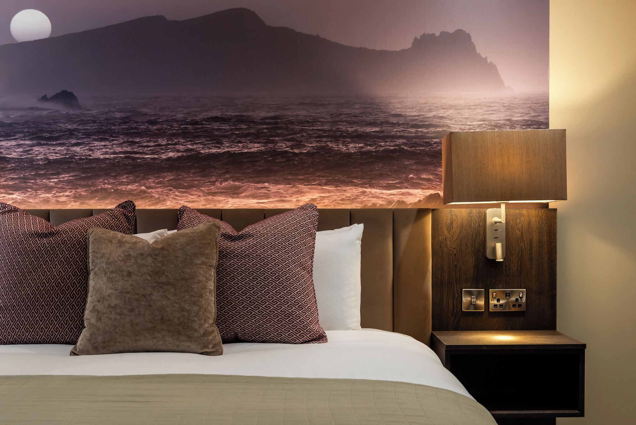 A bed with patterned pillows, a nightstand with a lamp, and a wall mural of a coastal scene.