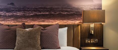 Una cama con almohadillas estampadas, una mesita de noche con lámpara y un mural en la pared con un paisaje costero.