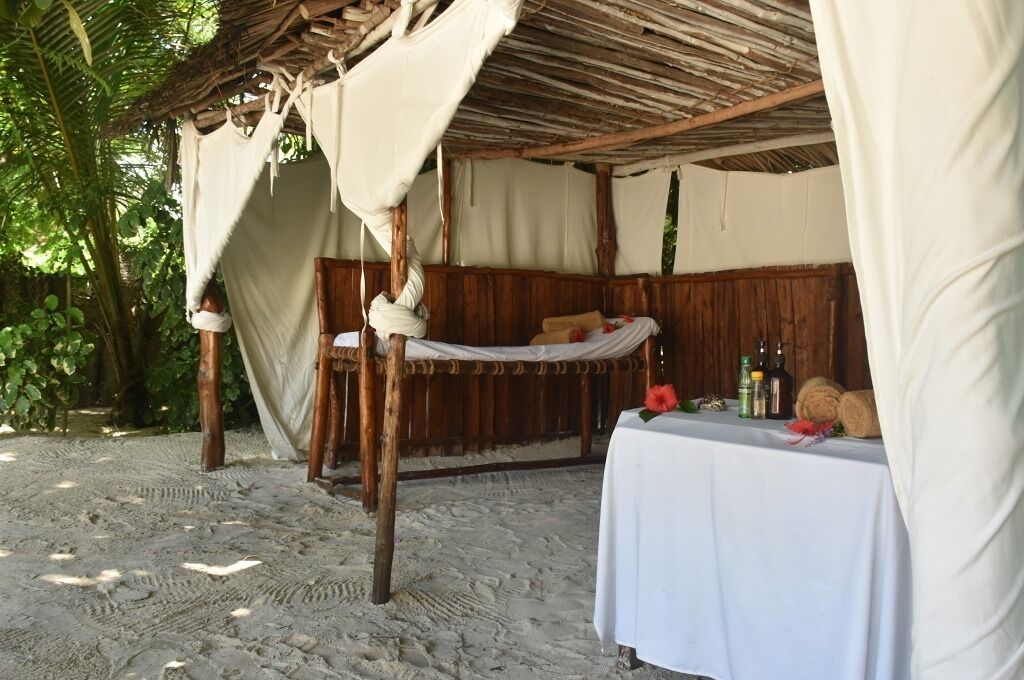 Une cabane rustique en bois, avec un toit de chaume, un hamac et une table sur laquelle se trouvent des bouteilles et une fleur.