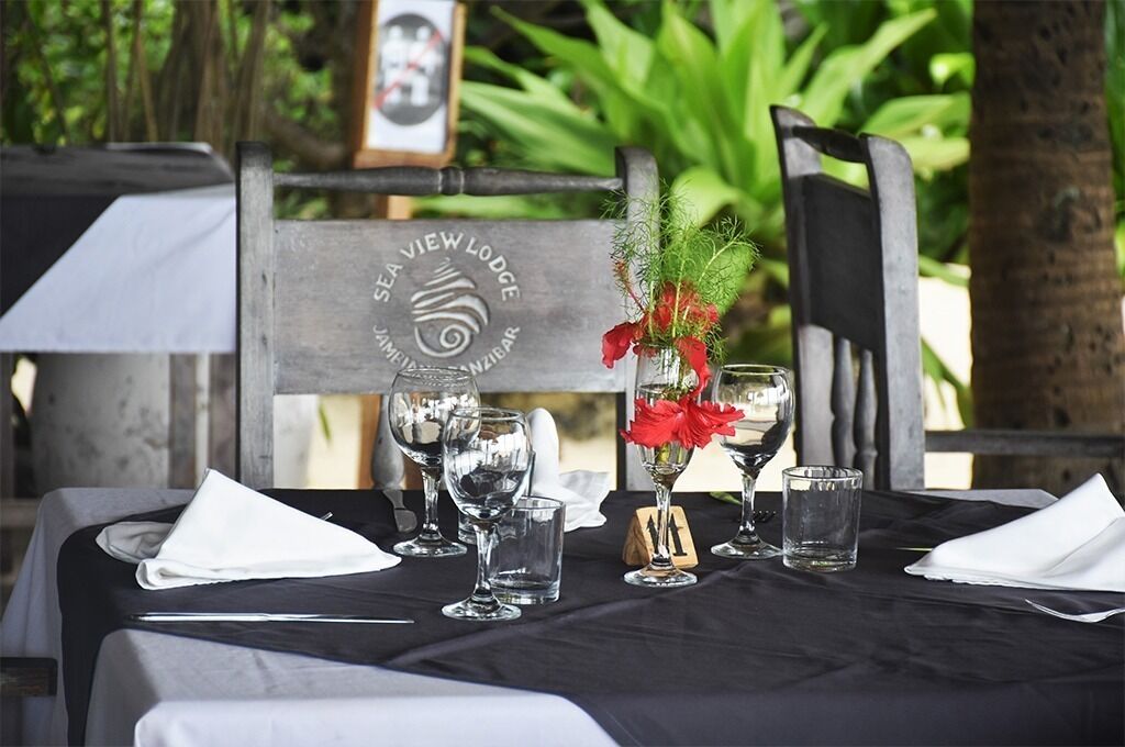 Une table dressée avec des verres à vin, un vase rempli de fleurs rouges et une chaise en bois portant une plaque avec l’inscription « SEA VIEW LODGE ».