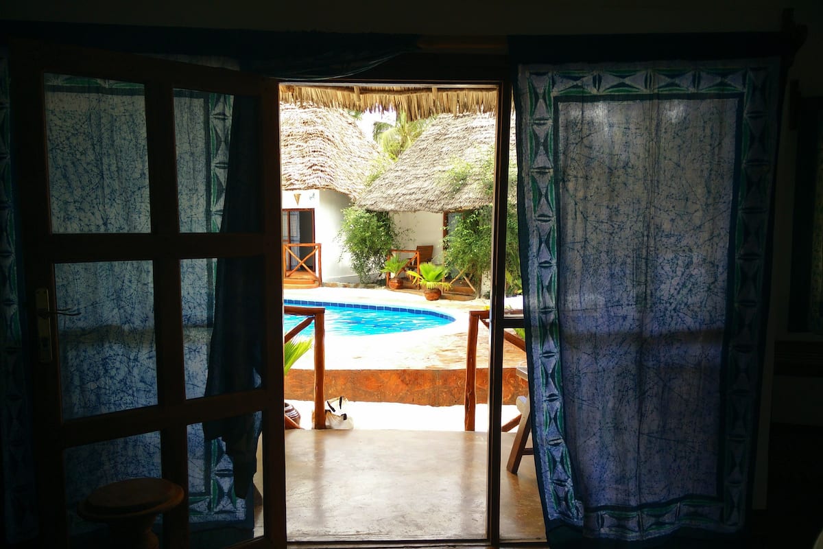 An open door leading to a pool area with a thatched roof and potted plants.