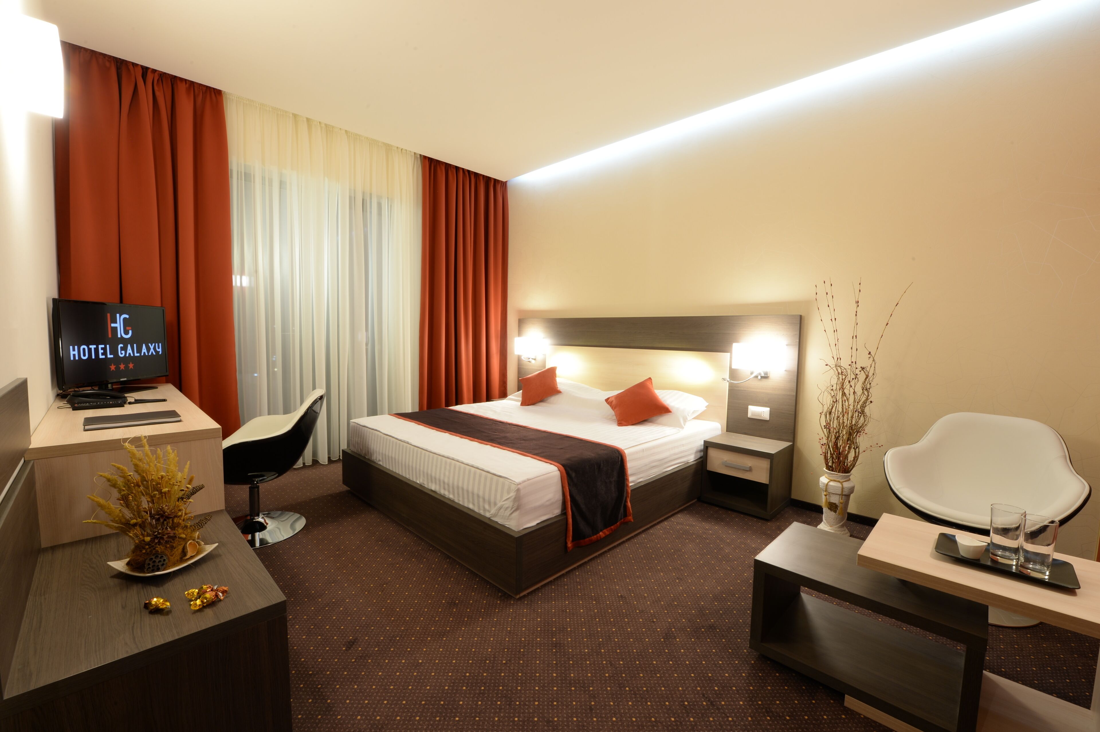 A hotel room with a bed, desk, chair, TV, bedside table, and a vase with branches.
