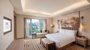 A hotel room with a large bed, a desk, a chair, a bench, a nightstand, a lamp, and a painting on the wall.