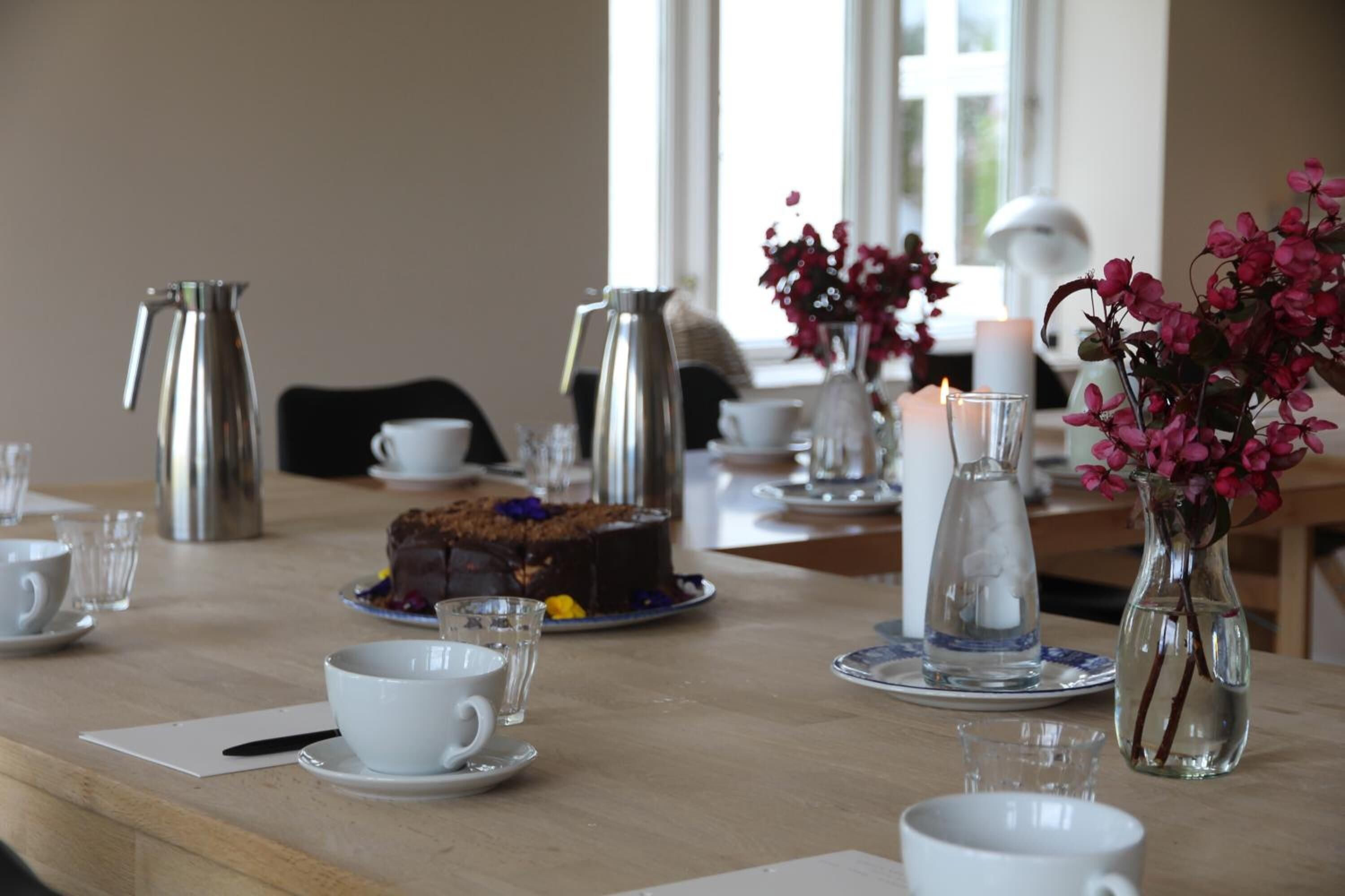 A dining table set with white cups, saucers, a cake, and a vase with pink flowers.