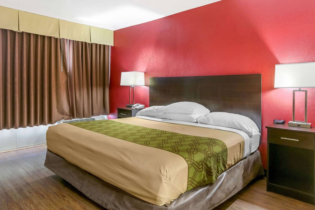 Una habitación de hotel con una cama, lámparas de noche, una mesita de noche, un aparador y una ventana con cortinas.