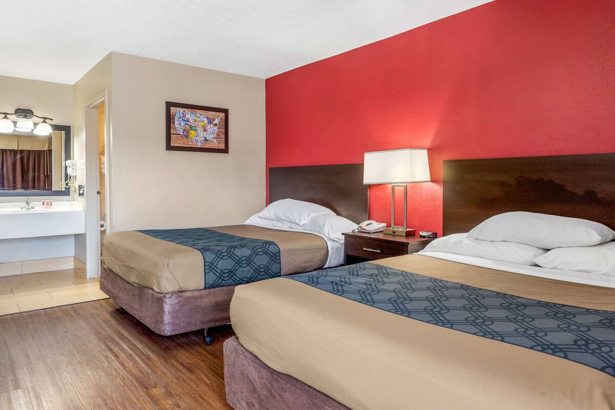 Habitación de hotel con dos camas, una pared de acento roja, suelo de madera, baño con espejo y lavamanos, y un cuadro en la pared.