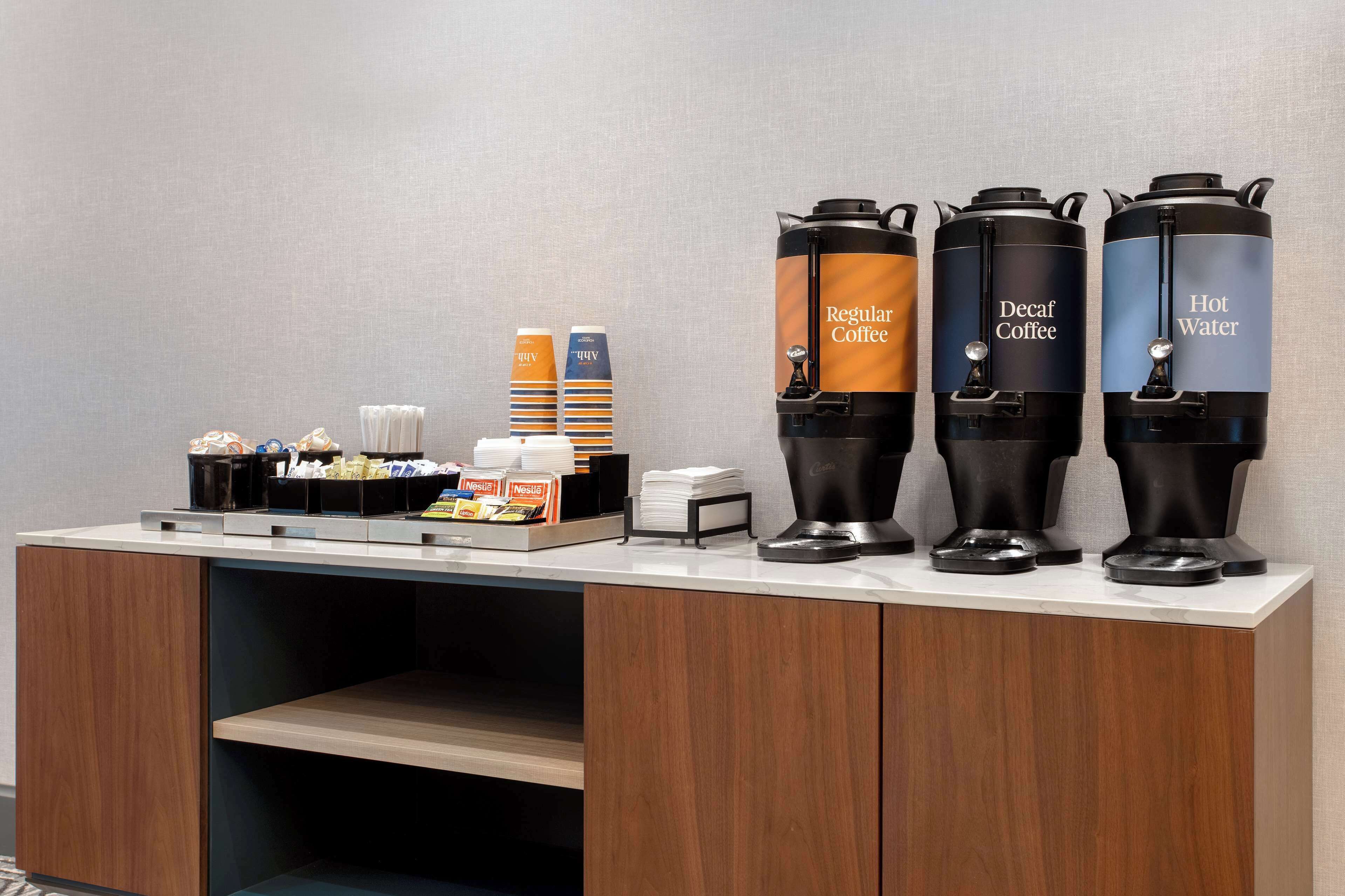 Estación de café con dispensadores para café regular, café descafeinado y agua caliente, además de una variedad de cápsulas de café y una pequeña pila de servilletas.