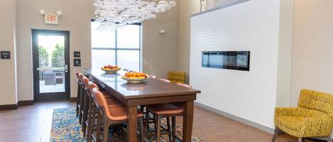 A modern dining area with a long table, chairs, a fruit bowl, a patterned rug, and a unique ceiling light fixture.