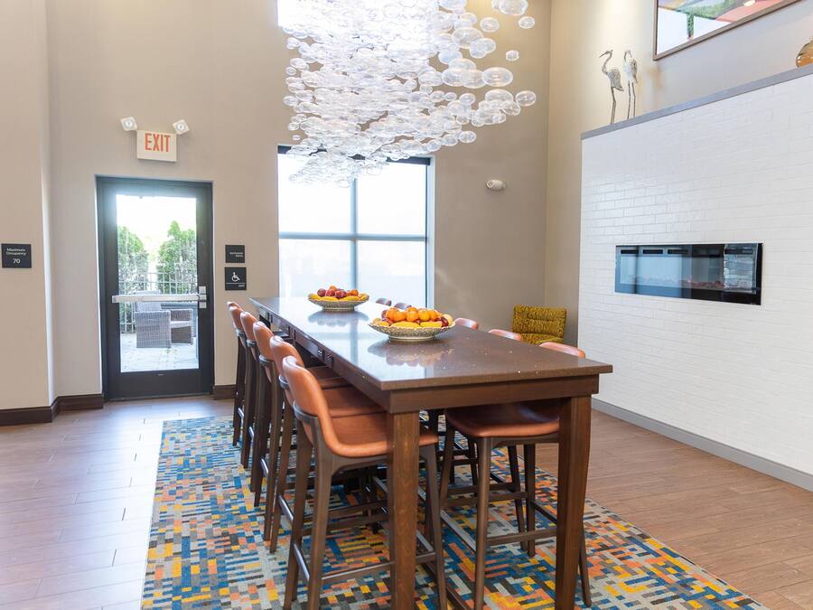 A modern dining area with a long table, chairs, a fruit bowl, a patterned rug, and a unique ceiling light fixture.