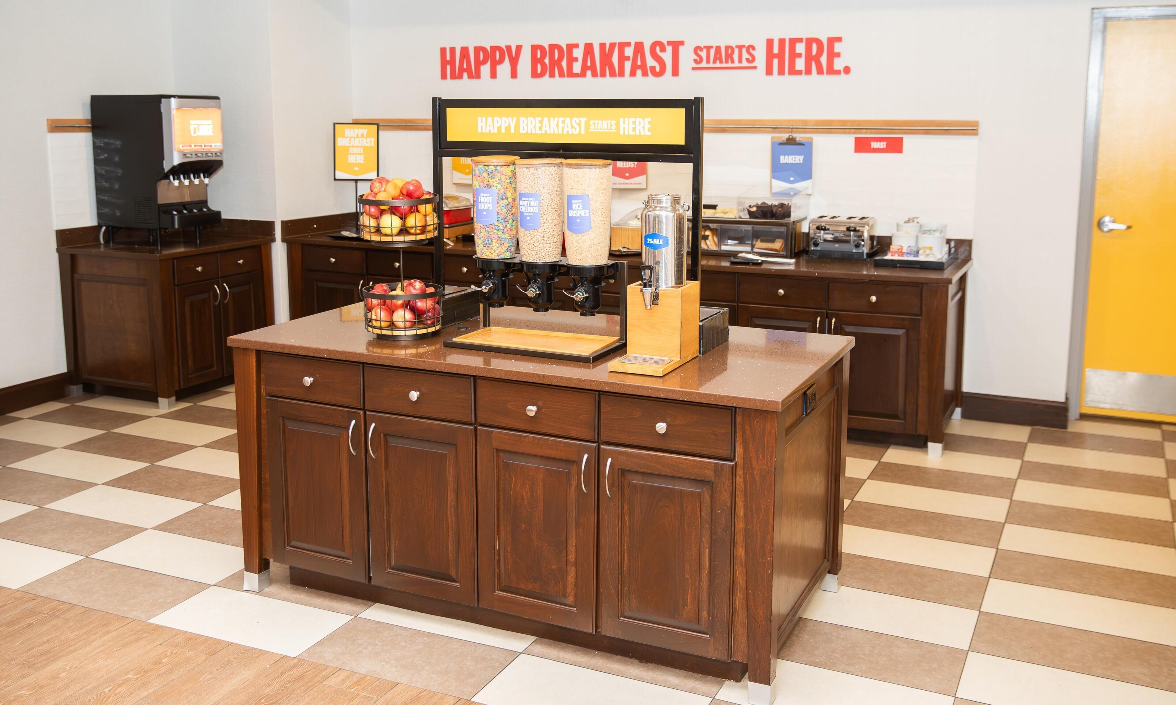 A breakfast bar with a sign reading 'HAPPY BREAKFAST STARTS HERE.' There are various food dispensers and a fruit bowl on the counter.