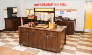 A breakfast bar with a sign reading 'HAPPY BREAKFAST STARTS HERE.' There are various food dispensers and a fruit bowl on the counter. - Hampton Inn & Suites Williamsport-Faxon Exit (Williamsport)