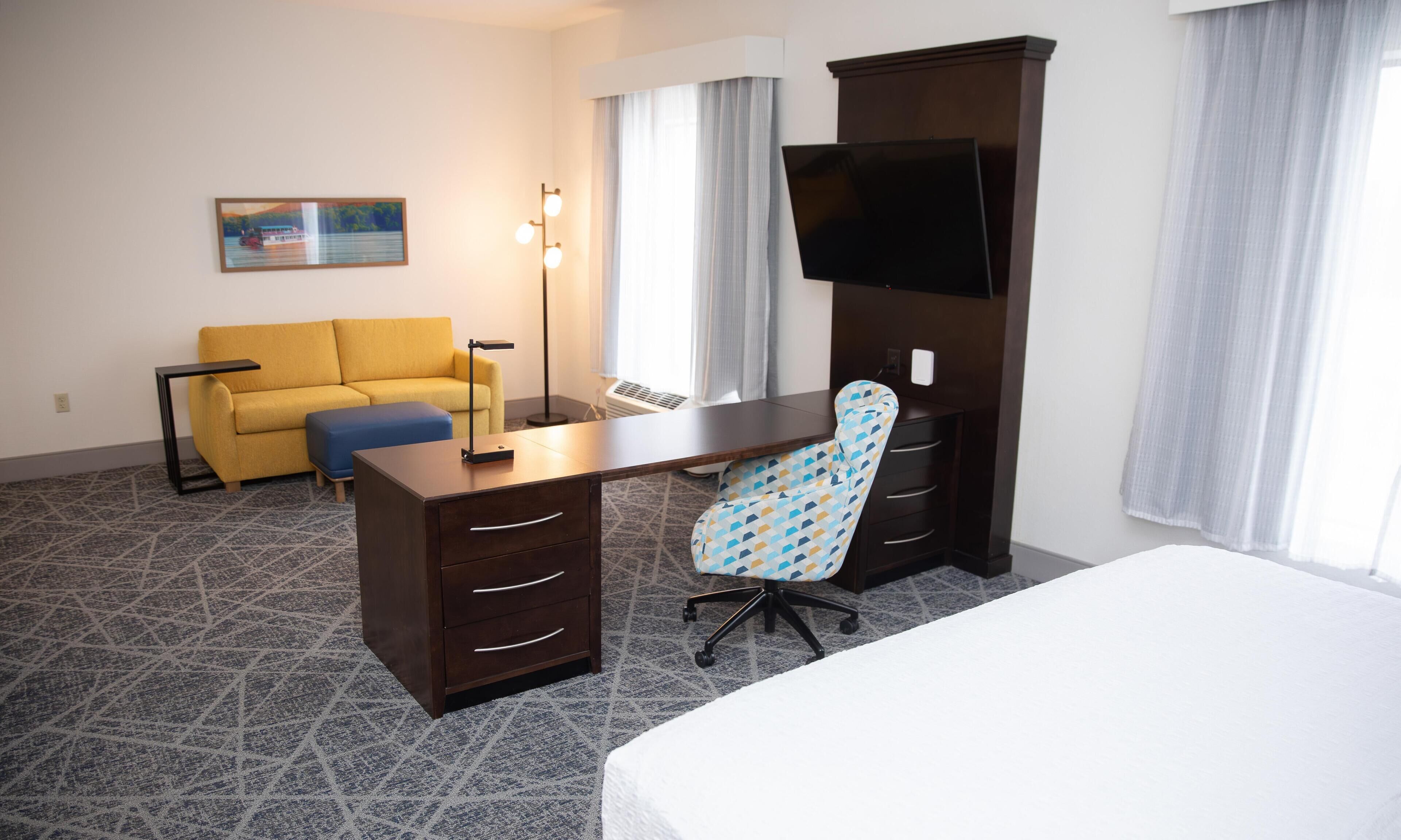 A hotel room with a bed, a desk with drawers, a chair, a sofa, a TV, and a painting on the wall.