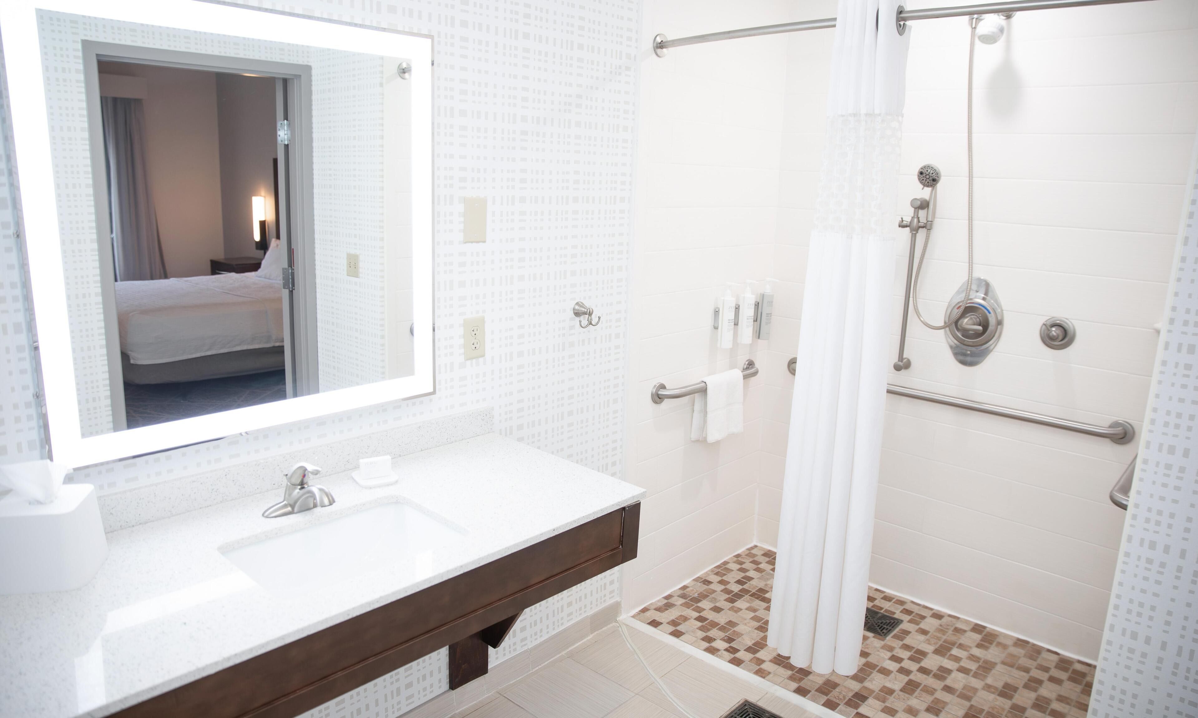 A bathroom with a large mirror, a sink, a shower with grab bars, and a tiled floor.
