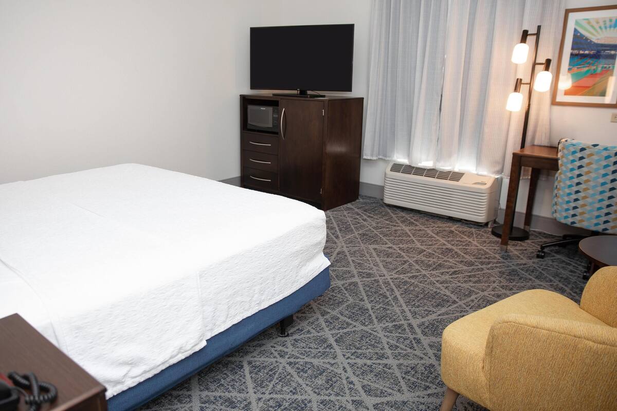 A hotel room with a bed, a desk, a chair, a TV, a heater, and a window with curtains.