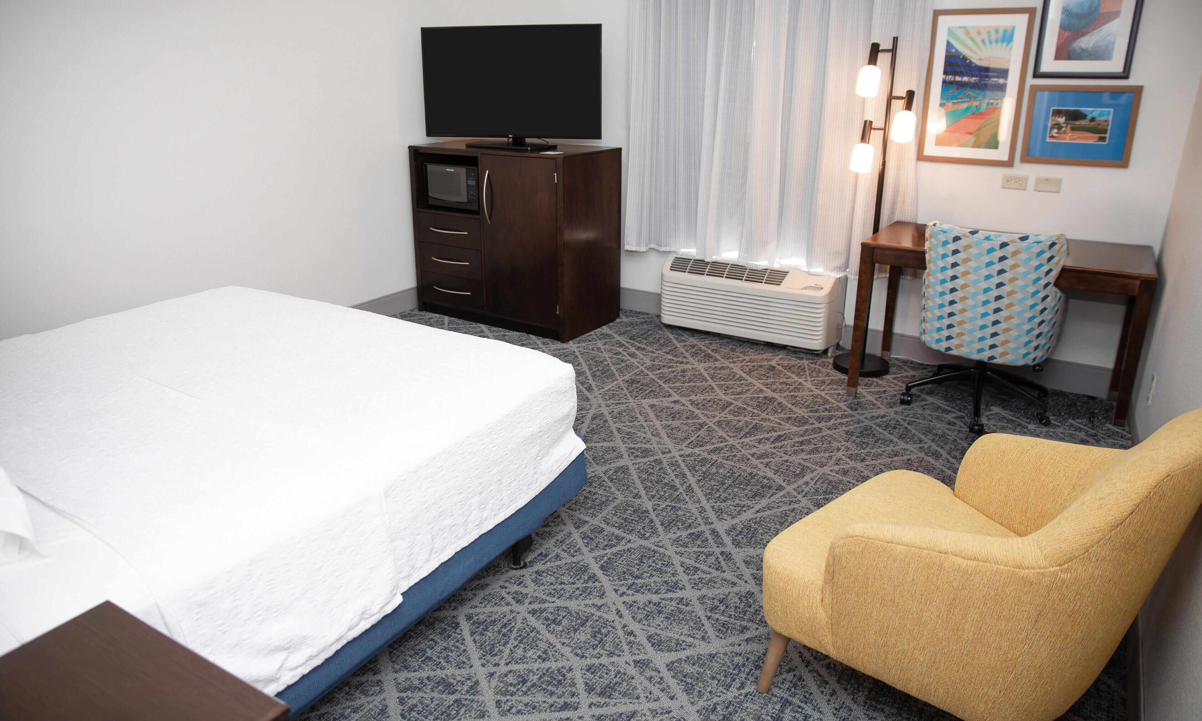 Una habitación de hotel con una cama, un escritorio con silla, un televisor, un calentador y cuadros en la pared.