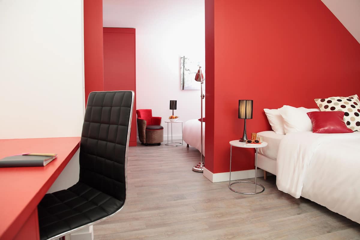Une chambre d’hôtel moderne avec un mur d’accent rouge, un fauteuil de bureau noir, un lit blanc avec des oreillers rouges et blancs, une petite table ronde et un lampadaire.