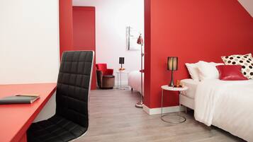 Una habitación de hotel moderna con una pared de acento roja, una silla de oficina negra, una cama blanca con almohadas rojas y blancas, una mesita redonda pequeña y una lámpara de pie.