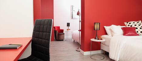 A modern hotel room with a red accent wall, a black office chair, a white bed with red and white pillows, a small round table, and a floor lamp.