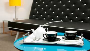 Een moderne kamer met een zwart gestoffeerde bank, een felgele lamp en een blauwe dienblad met een witte theepot, twee zwarte kopjes en een lepel.