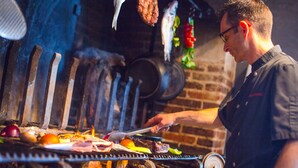 Un cocinero asando carne y verduras en una parrilla, con un muro de ladrillo al fondo.