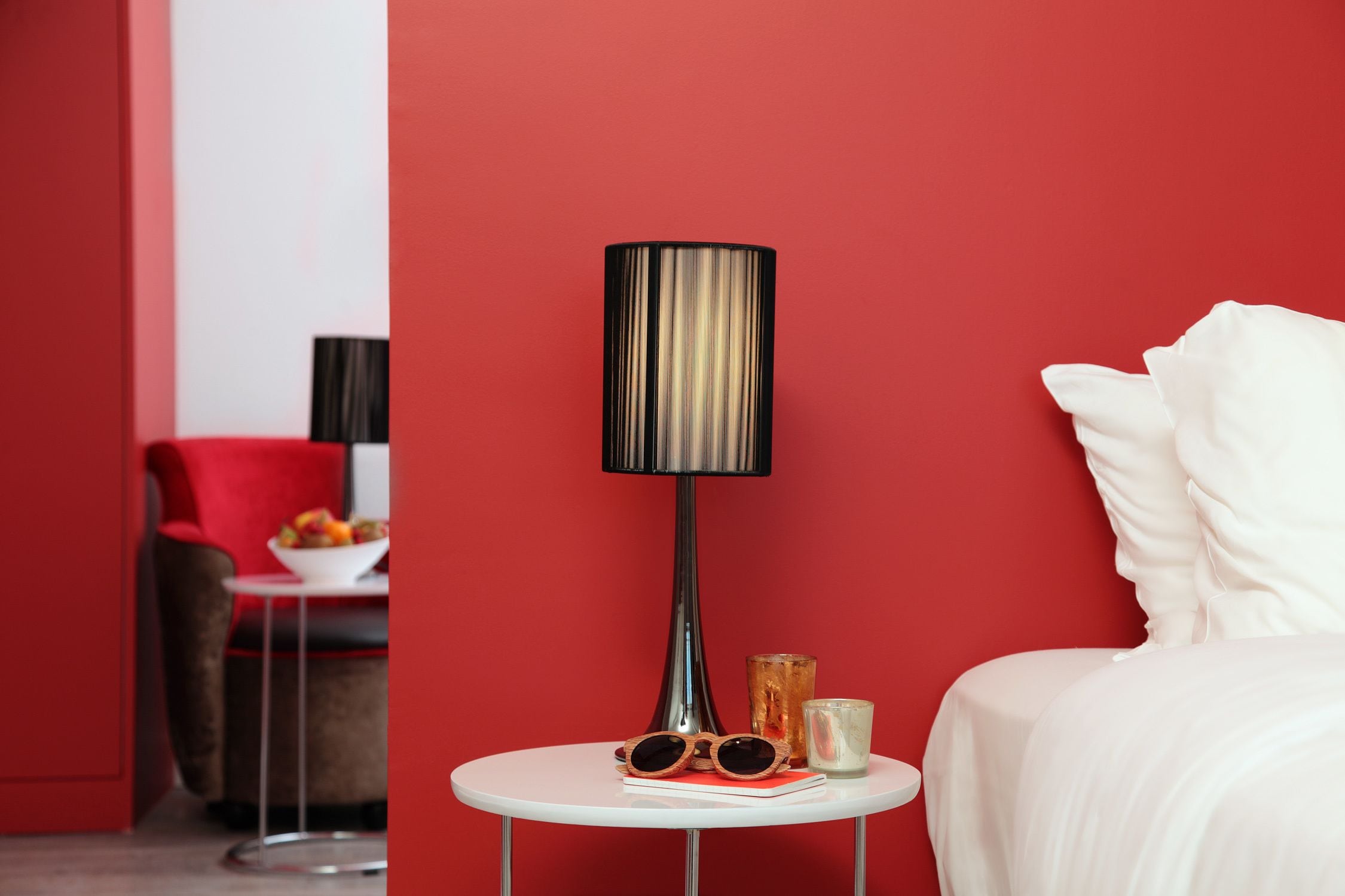 Una habitación de hotel moderna con una pared roja, una cama blanca, una mesa redonda blanca con una lámpara, gafas de sol y un vaso.