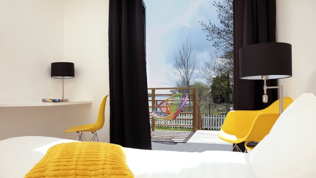 Habitación de hotel con cama, una silla amarilla, una hamaca y vistas al exterior.