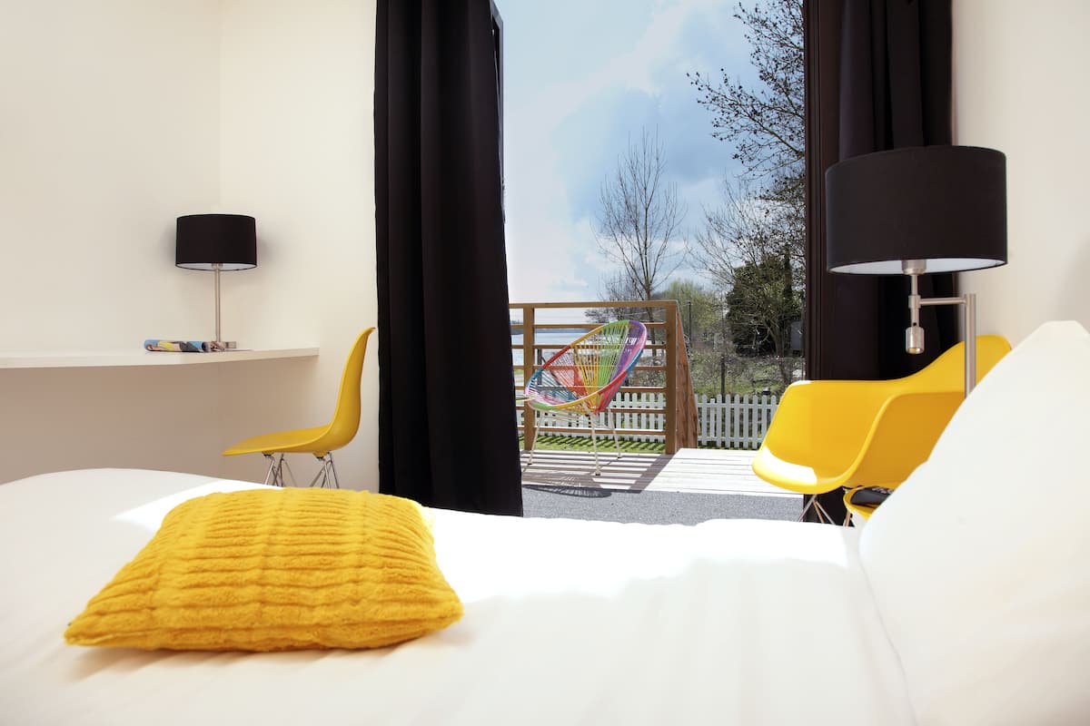 Une chambre d’hôtel avec un lit, une chaise jaune, un hamac et une vue sur l’extérieur.