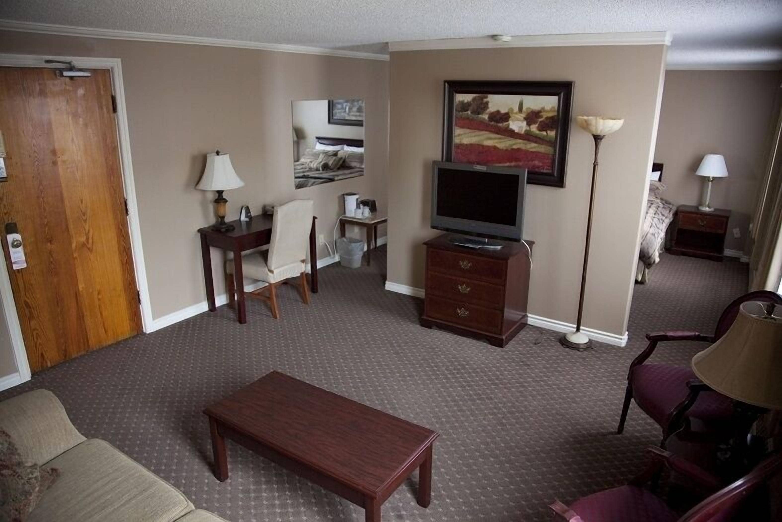 Une chambre d’hôtel avec un canapé en bois, une table basse en bois, un téléviseur posé sur un meuble, un bureau avec une chaise, une lampe et un tableau accroché au mur.