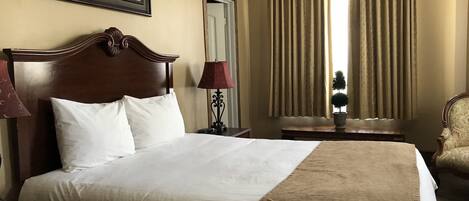 Habitación de hotel con una cama grande, una mesita de noche, una lámpara, una silla, una ventana con cortinas y un cuadro en la pared.