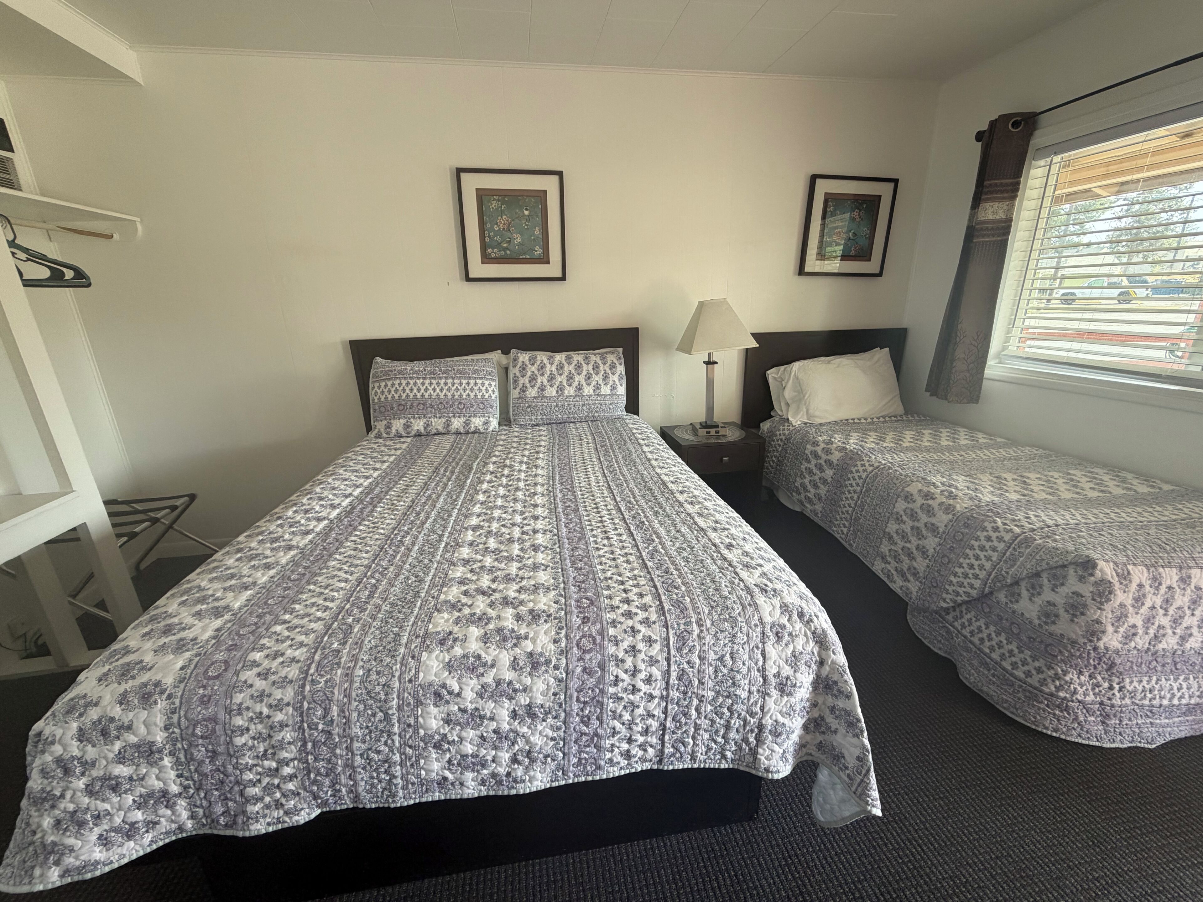 A hotel room with two beds, a nightstand, a lamp, and two framed pictures on the wall.