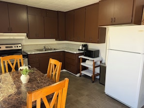A kitchen with dark brown cabinets, a white refrigerator, a microwave on a small shelf, a stove, and a dining table with chairs. - Sonoma Lodge (Bend)