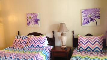 A hotel room with two beds, each with a colorful zigzag patterned bedspread. There are two wall-mounted lamps between the beds, and two wall-mounted flower paintings.