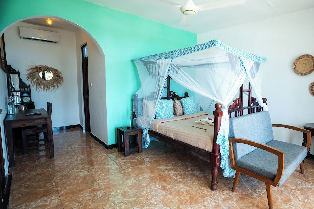 A room with a four-poster bed, a wooden chair, a desk with a chair, and a television on the wall.