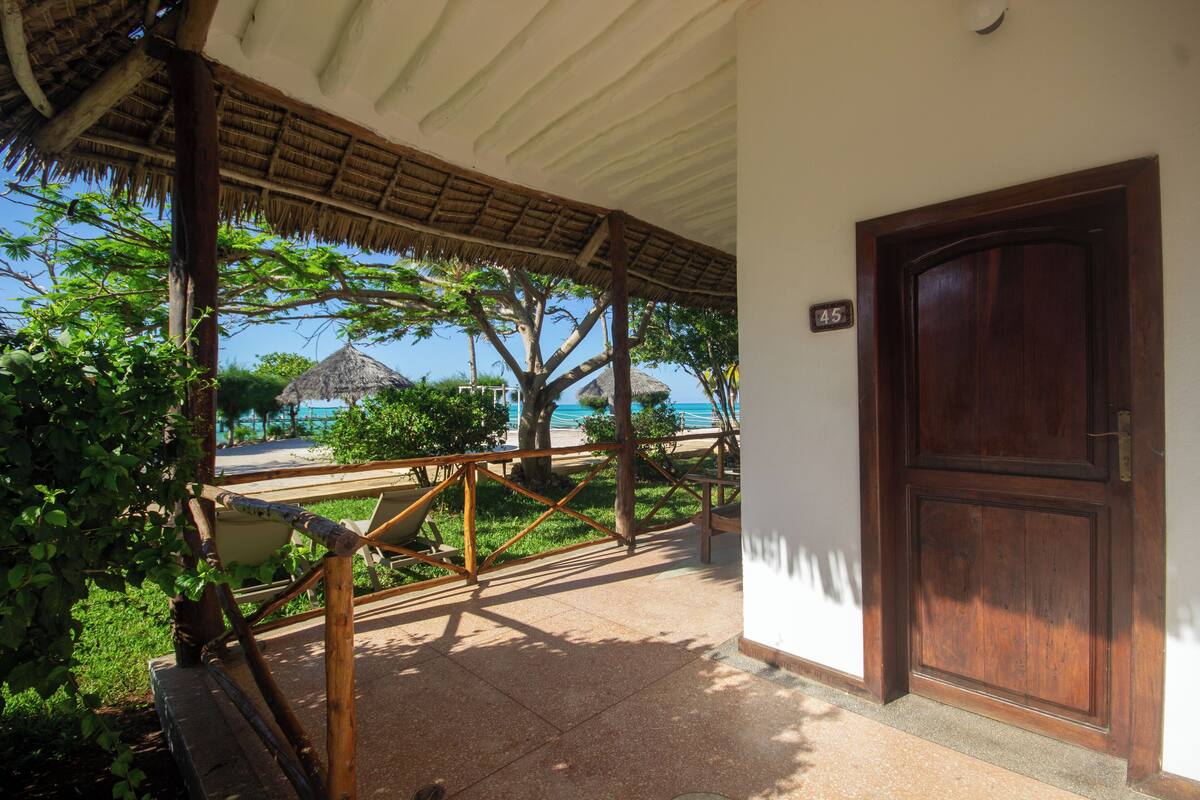 A wooden door with a number plate, a covered porch with a thatched roof, and a view of the beach and ocean.