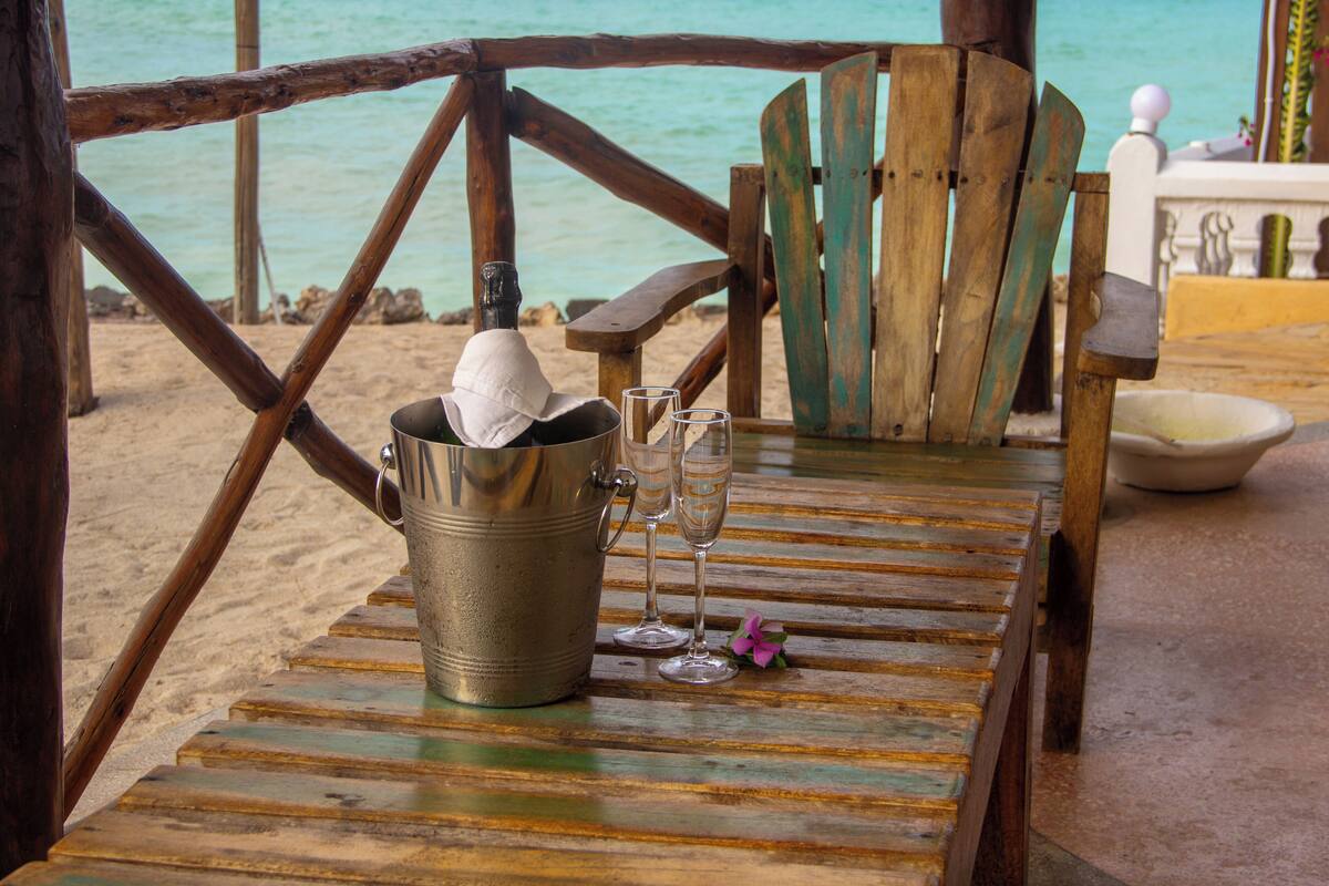 A wooden table with a metal bucket, two wine glasses, and a bottle of champagne on a beach setting.