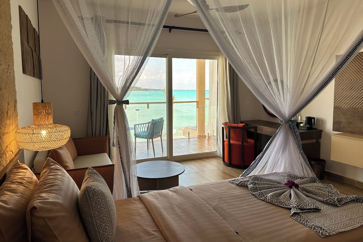 A bedroom with a bed, a bedside table, a chair, and a view of the ocean through a large window.