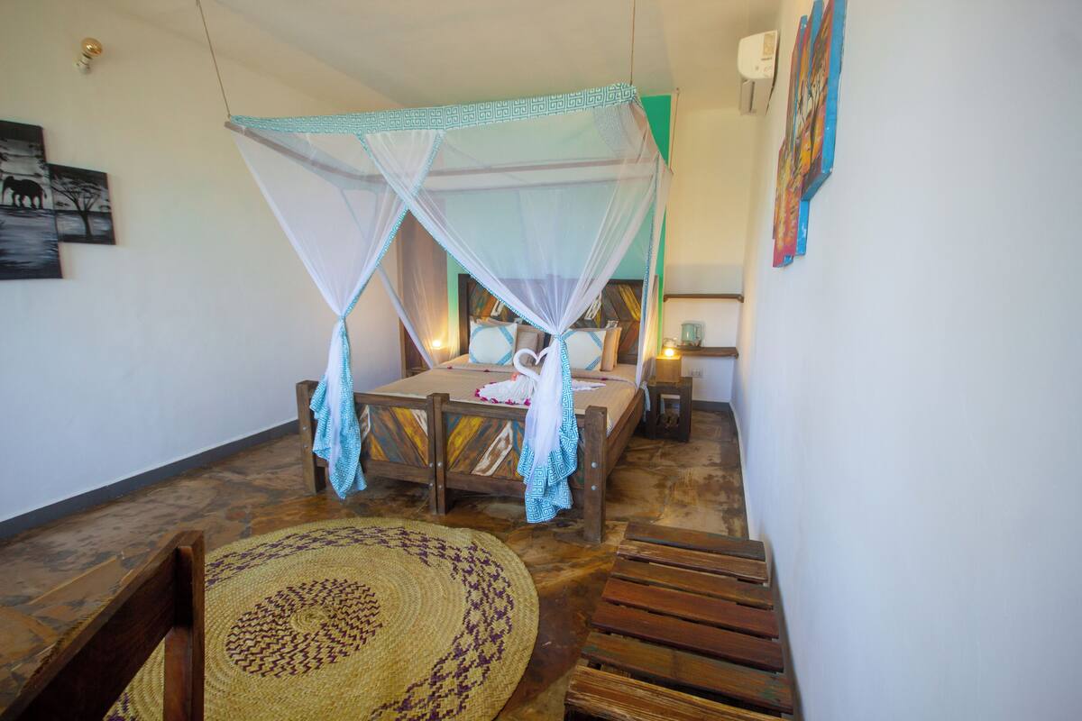 A room with a bed under a mosquito net, a wooden chair, a woven rug, and a wall-mounted air conditioner.
